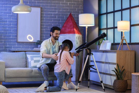 Little girl with her father looking through telescope at home in evening