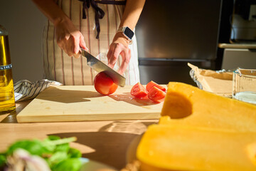 Close-up of woman cutting tomato on wooden board in kitchen with vegetables and pumpkin on table. Concept of organic food and healthy cooking