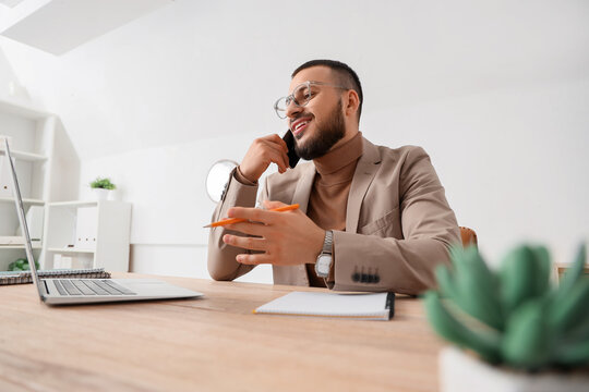 Young businessman with laptop talking by mobile phone in office