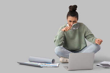 Young woman with magnifier and laptop sitting on grey background. Search concept