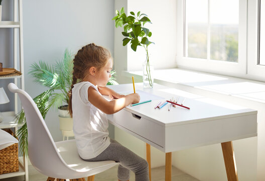 Little preschool girl sitting at table in front of big window, drawing with colorful pencils. Focused kid painting picture in bright room, developing creativity. Education, art, homeschooling concept.