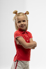 little blonde girl stands in a studio crossing her arms with a smile. She wears a red shirt and shorts showcasing her playful spirit in a bright setting.