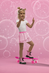 small blonde girl stands confidently on a pink skateboard in a studio setting. She is wearing a white shirt pink skirt and boots. Bubbles surround her in the playful scene.