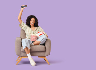 Female African-American sports fan with popcorn in armchair watching football on lilac background