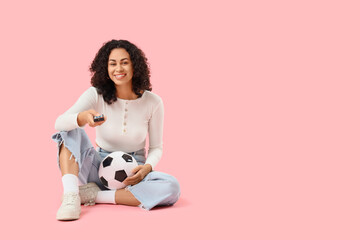 Female African-American sports fan with ball watching soccer on pink background