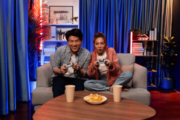 Sitting sofa of happy smiling couple playing video game control by joystick fronted snack and drinks competing challenge level with funny at modern neon blub light comfy living room at home. Infobahn.