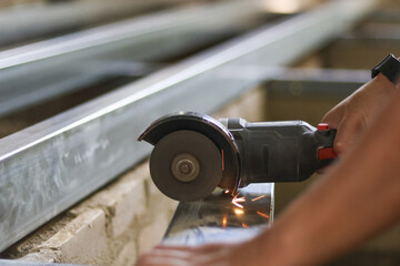 Focused worker using power tool, an angle grinder, to cut metal steel stud at construction site. Sparks fly from cutting wheel, showing industrial fabrication process