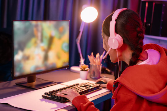 Host channel of young beautiful Asian gaming streamer playing fighting Moba at battle arena game on pc screen with multiplays, wearing pastel headphone at neon digital light modern room. Stratagem.