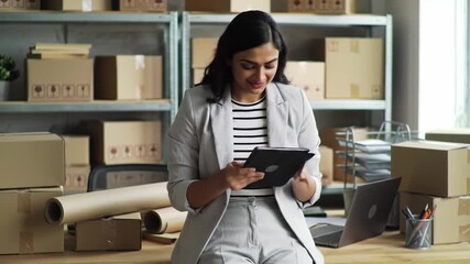 Woman smiles using a tablet in a warehouse office with shelves and boxes. She wears a striped shirt and gray blazer, with a laptop and desk nearby. - Powered by Adobe