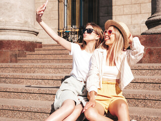 Two young beautiful smiling hipster female in trendy summer clothes.carefree women posing in the street. Positive pure models having fun at sunset, hugging and taking selfie.Sitting at the stairs