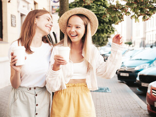 Two young beautiful smiling hipster female in trendy summer clothes.Sexy carefree women posing in the street. Positive pure models having fun at sunset. They drinking coffee or tea in plastic cup