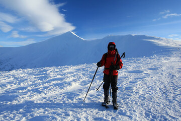 A winter mountaineer in a red jacket stands on a snowy ridge with trekking poles. Bright cold landscape, blue sky and vast mountains create a feeling of harsh high-altitude adventure.