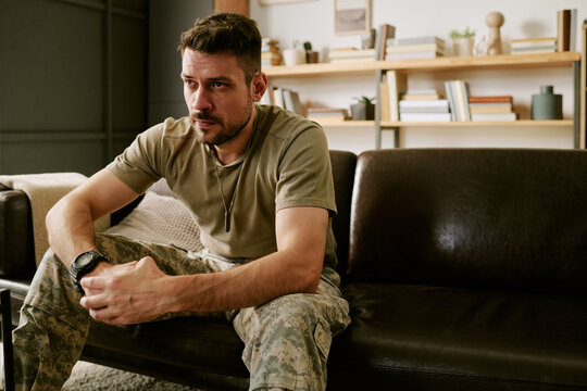 Caucasian young adult man sitting on couch leaning forward with hands clasped looking thoughtful wearing military camouflage pants representing psychological stress or PTSD - Powered by Adobe