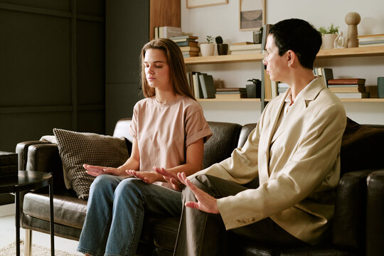 Young Caucasian woman practicing mindfulness exercise with middle aged Caucasian woman guiding session on couch in modern office setting, both focusing on relaxation techniques