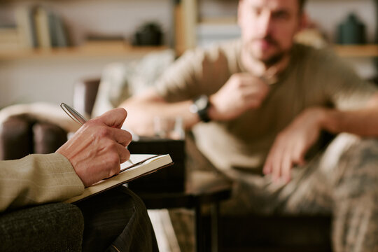 Caucasian middle aged woman holding pen and writing in notebook while listening to blurred Caucasian middle aged man talking during psychological counseling session