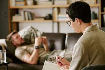 Middle aged Caucasian man lying on couch talking, while middle aged woman psychologist sitting in foreground writing notes during therapy session in office setting