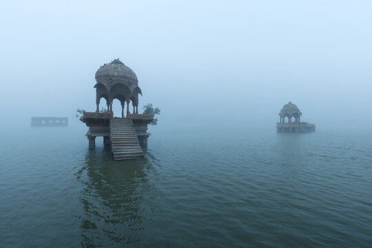 Misty dawn view of Gadisar Lake in Jaisalmer, Rajasthan, with ornate sandstone chhatris emerging from the mist above calm rippling water, creating a serene and mystical atmosphere.
