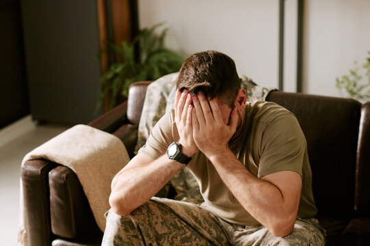 Caucasian young adult man in military uniform sitting on couch covering face with hands showing signs of emotional distress highlighting psychological struggle in soldier