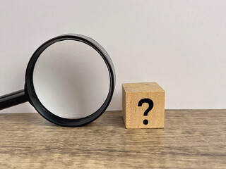 Question mark written on wooden block with magnifying glass next to it on wooden background.