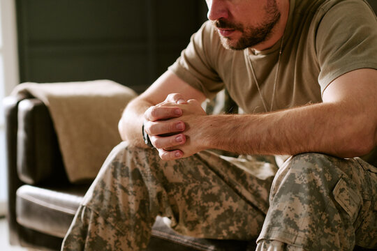 Caucasian middle aged man sitting on couch clasping hands together appearing deep in thought wearing military uniform representing psychological stress or contemplation
