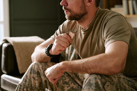 Caucasian young adult man sitting indoors holding military dog tags in hand appearing thoughtful, wearing camouflage pants, showing close up of face and upper body, psychology concept