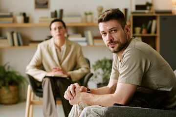 Portrait of Caucasian young adult man sitting in therapy session looking at camera with serious expression, middle aged Caucasian woman psychologist sitting in background taking notes