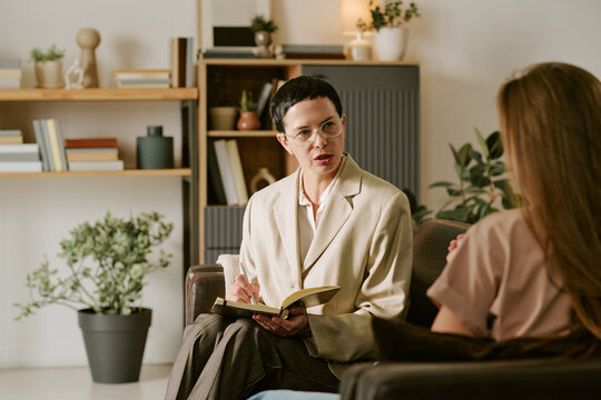 Middle aged Caucasian woman therapist listening attentively and taking notes while young adult Caucasian woman client sitting on couch during psychology counseling session