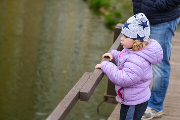 Little girl in lavender jacket with stars hat standing at wooden railing above water. Childhood outdoor activity and exploration concept.