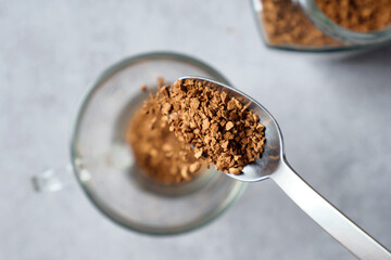 Freeze-dried instant coffee beans falling from a metal teaspoon into an empty cup on background of table with coffee glass jar.