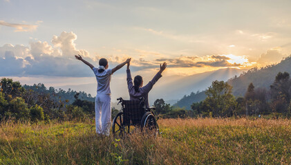 Silhouette elderly woman in wheelchair raised hands with his care helper on mountain sunset.
