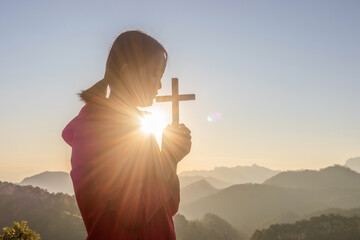 Girl praying and holding Christian cross for worshipping God on mountain at sunrise background. Christian, Christianity, Religion copy space background. Easter Sunday concept: