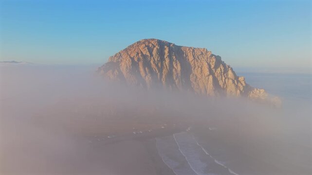 Aerial view shows a stunning Morro rock formation rising above fog, revealing a peaceful coastal landscape at dawn, showcasing natures beauty during the enchanting sunrise. Morro Bay, California