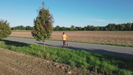 Man jogging on empty country road near agricultural field and green trees on sunny day. Athlete training outdoors. Morning running. Concept of fitness, outdoor activity and healthy lifestyle