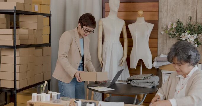 Crafting Confidence: A small business owner meticulously organizes merchandise in her workshop, while a seasoned seamstress offers her support.