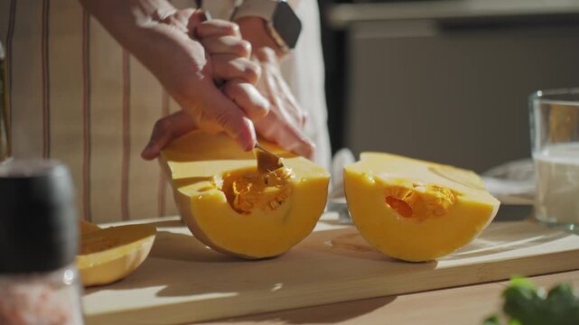 Close-up of woman removing seeds from halved pumpkin with spoon on wooden cutting board in kitchen. Person in apron preparing squash for roasting. Concept of home cooking and healthy food