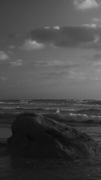 Dramatic black-and-white seascape with strong contrast, showing a large coastal rock in the foreground and dynamic waves under a cloudy sky. The monochrome tones emphasize texture, atmosphere