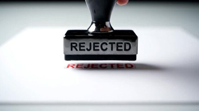Hand presses a "REJECTED" stamp onto a white surface, close-up, angled shot