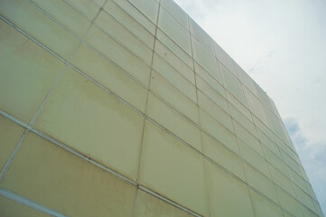 Low angle view of a large, modern building with a pale yellow facade, featuring a grid pattern and visible seams against a cloudy sky, creating an architectural perspective.