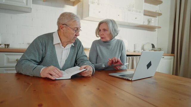 Elderly people spending weekend at home, woman using gadgets, man reading book. Portrait of senior married couple in kitchen, grandfather and grandmother have different passion and favorite leisure