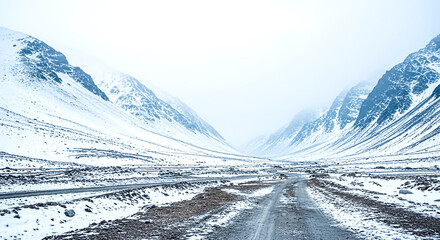 A winding road through a snow-blanketed valley, surrounded by towering mountains under a gray, overcast sky, evoking tranquility.