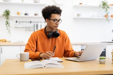 A young Black man wearing glasses and headphones works on a laptop at a wooden table, with a book and coffee cup nearby.
