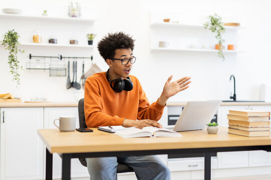 A young man with glasses is engaged in a video call, gesturing with his hand while seated at a table in a bright kitchen setting. - Powered by Adobe