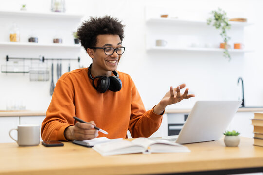 A young man with glasses smiles while participating in a video call on his laptop, gesturing with his hands.