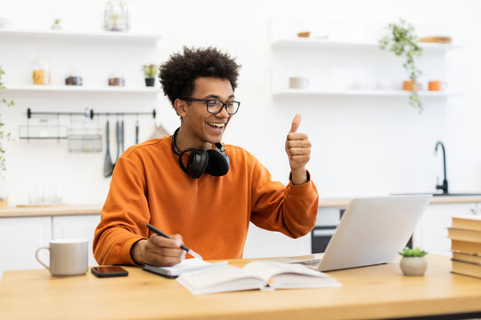 A young man with glasses gives a thumbs up while on a video call, working from home with a laptop and notebook.