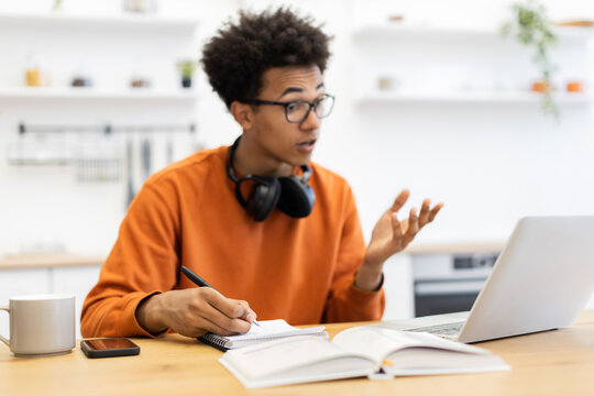 A young Black man with glasses is engaged in an online class, taking notes while looking at his laptop screen.