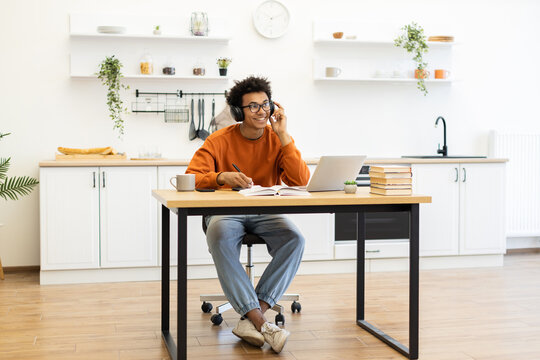 A young man with headphones smiles while working on a laptop at a table in his kitchen. - Powered by Adobe