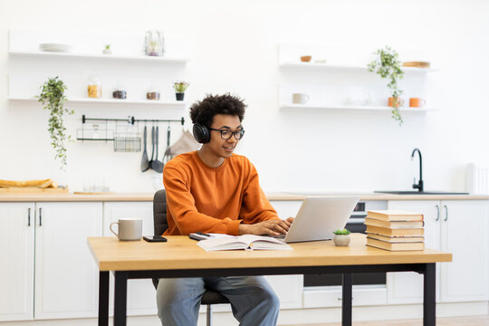 A young man with headphones works on a laptop at a table in a modern kitchen setting. - Powered by Adobe