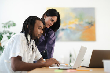 Diverse colleagues collaborate on a project in a modern office. The man types on a laptop while the woman observes and discusses the work. They are focused on their tasks.