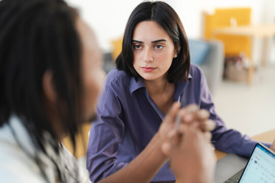A serious young woman in a purple shirt listens attentively to her male colleague during a professional discussion. They are working together in a bright office environment. - Powered by Adobe