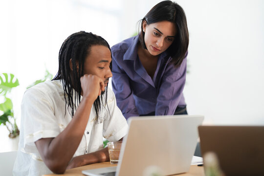 Two diverse colleagues collaborate on a project, reviewing content on a laptop screen. The man sits while the woman stands, both intently focused on their shared work.